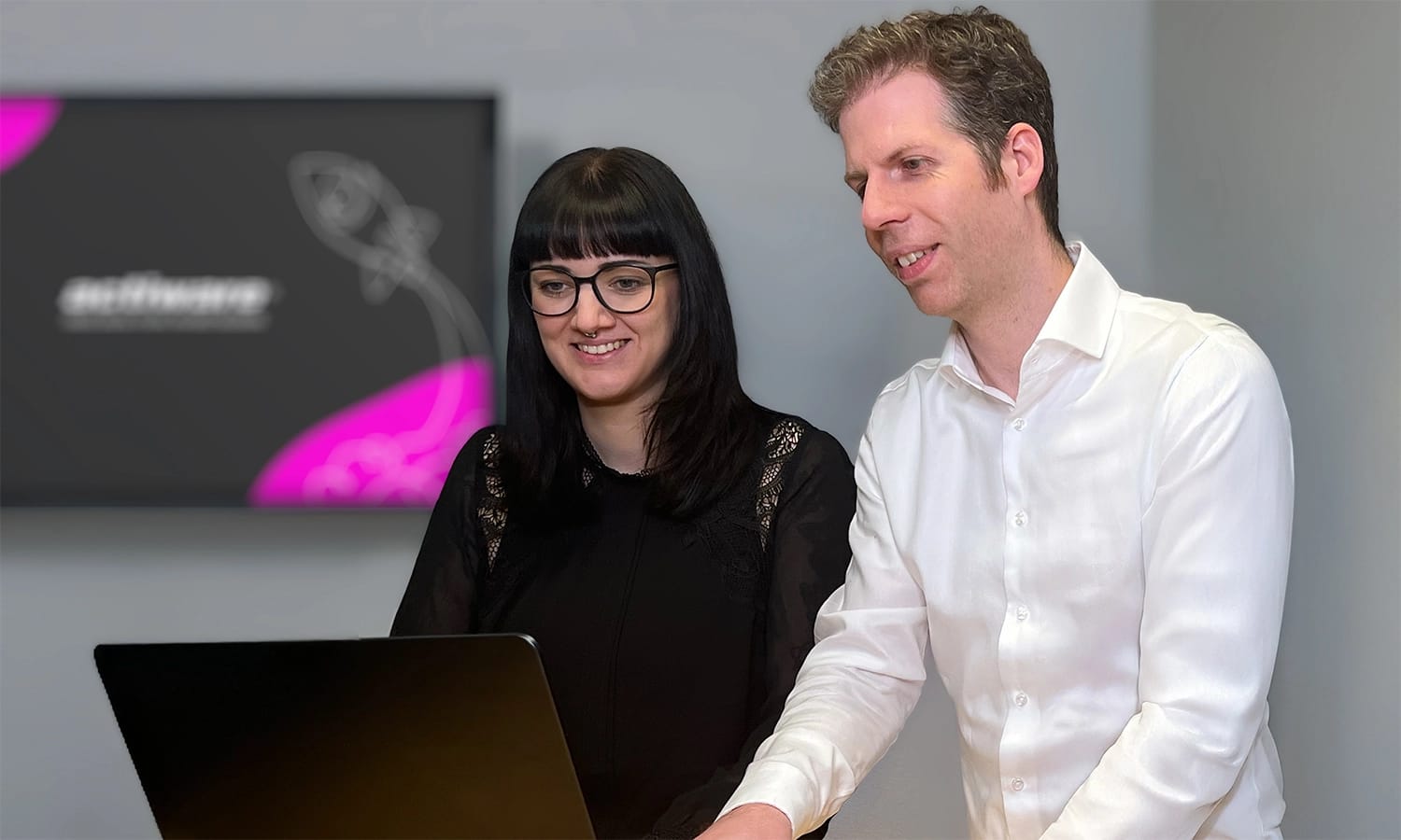 Zwei Mitarbeitende arbeiten zusammen, im Hintergrund hängt ein Bildschirm mit ACTIWARE-Branding.